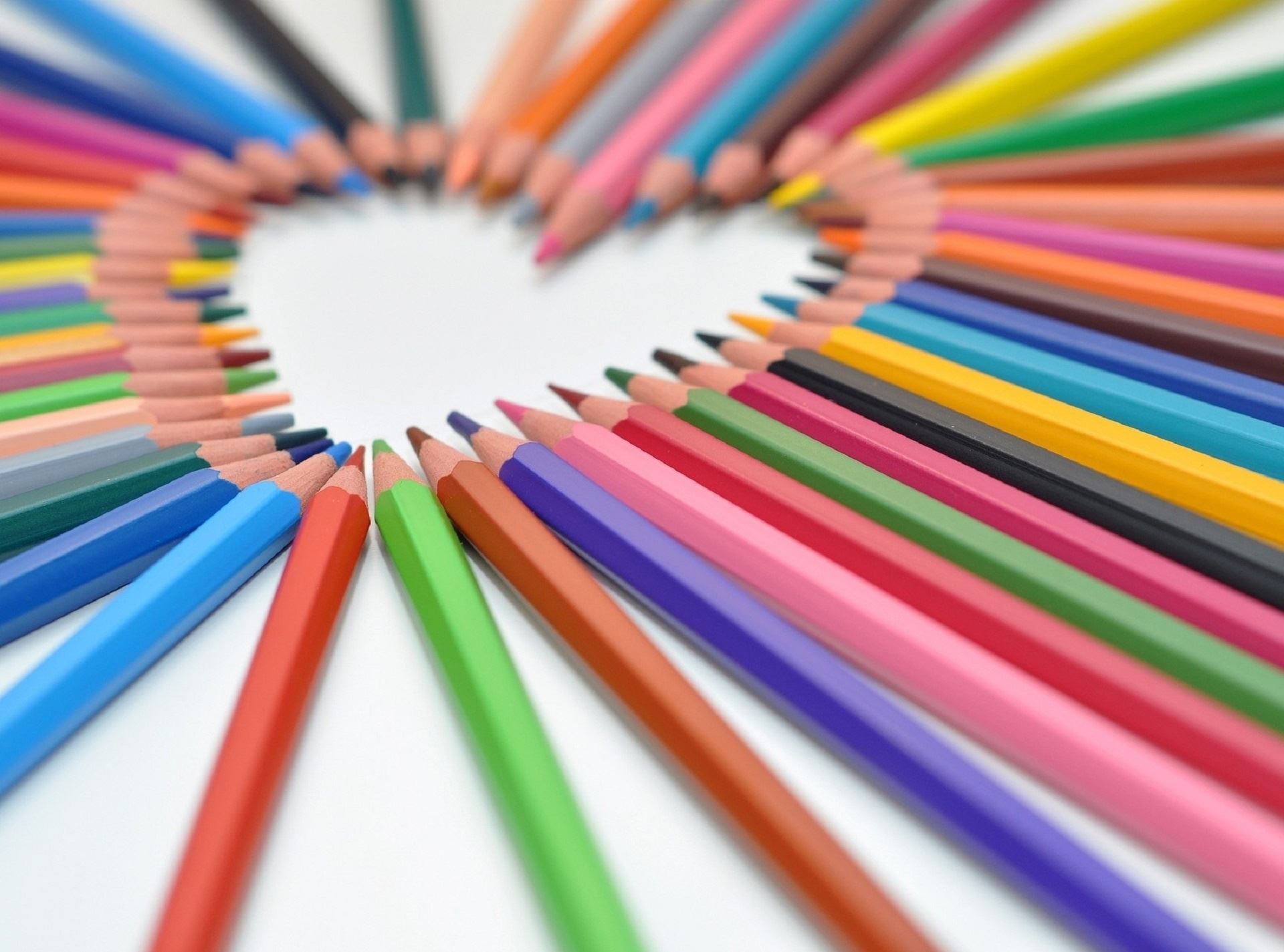 Rainbow color pencils laying on a white surface with their leads creating a heart-shaped open space. 